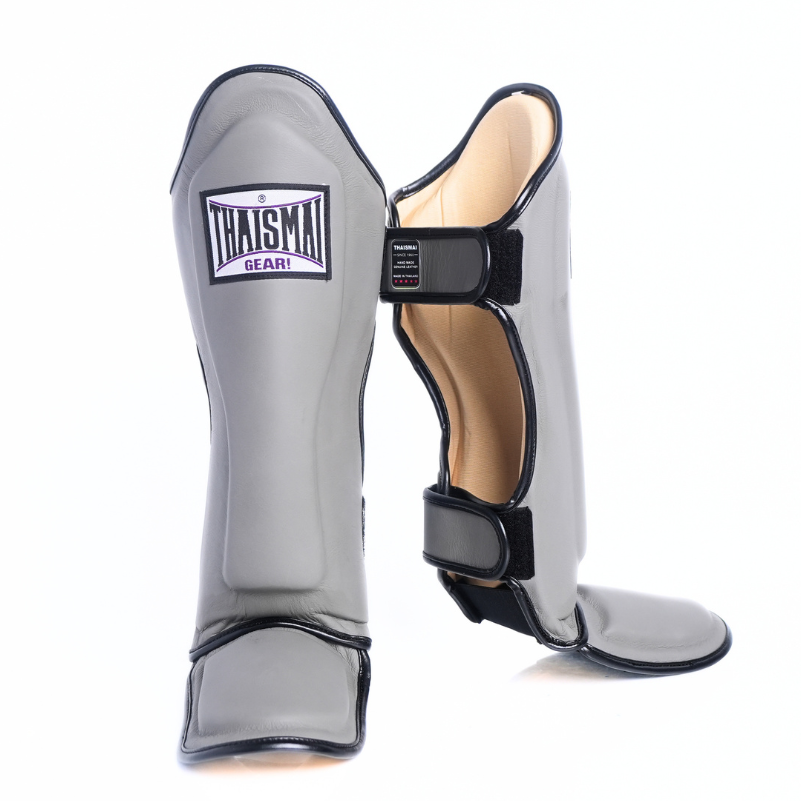 A pair of gray shin guards with black trim and hook-and-loop straps, shown on a clean white background. One guard is standing upright, showing the front with a 'THAISAI SHIN' brand patch, and the other is angled to show the side and back closure.