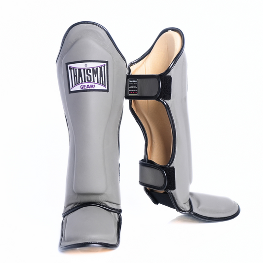 A pair of gray shin guards with black trim and hook-and-loop straps, shown on a clean white background. One guard is standing upright, showing the front with a 'THAISAI SHIN' brand patch, and the other is angled to show the side and back closure.