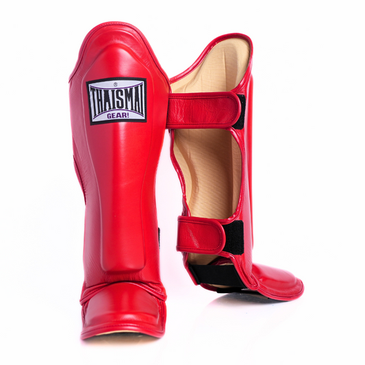 A pair of red shin guards for martial arts, with one standing upright to show the front and the other lying on its side to show the interior. The brand name 'THAISMA' is clearly visible on a white label on the upright shin guard.